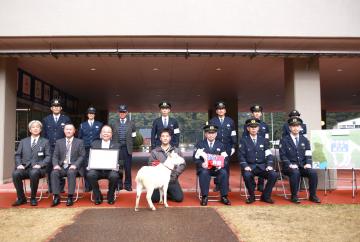 正装した警察職員や、市長、関係者と、中央で、リボンを付けたヤギと男性が並び記念撮影をしている写真