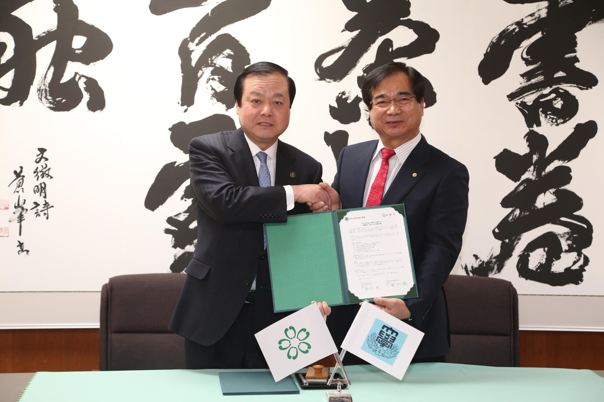 緑色のホルダーに協定書があり、そのホルダーを蕨市長と日本大学生産工学部 落合学部長と一緒に持ち片方の手で握手を交わしている写真