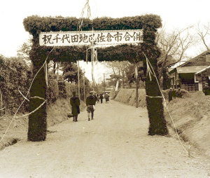 道路に「祝千代田地区佐倉市合併」と書かれたアーチの下を歩く人々の白黒写真