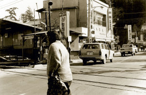 一人の女性が歩いている奥に踏切があり、その奥を車が走っている道路の白黒写真