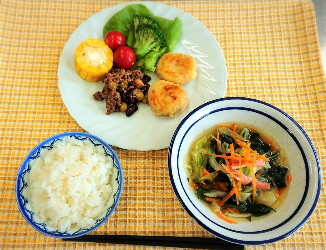 白いお皿に盛りつけられたツナ缶の焼きコロッケ、ミックスビーンズとひき肉の豆板醤炒め、付け合わせのコーン・プチトマト・ブロッコリーなどの野菜とコンソメ野菜スープとご飯の写真