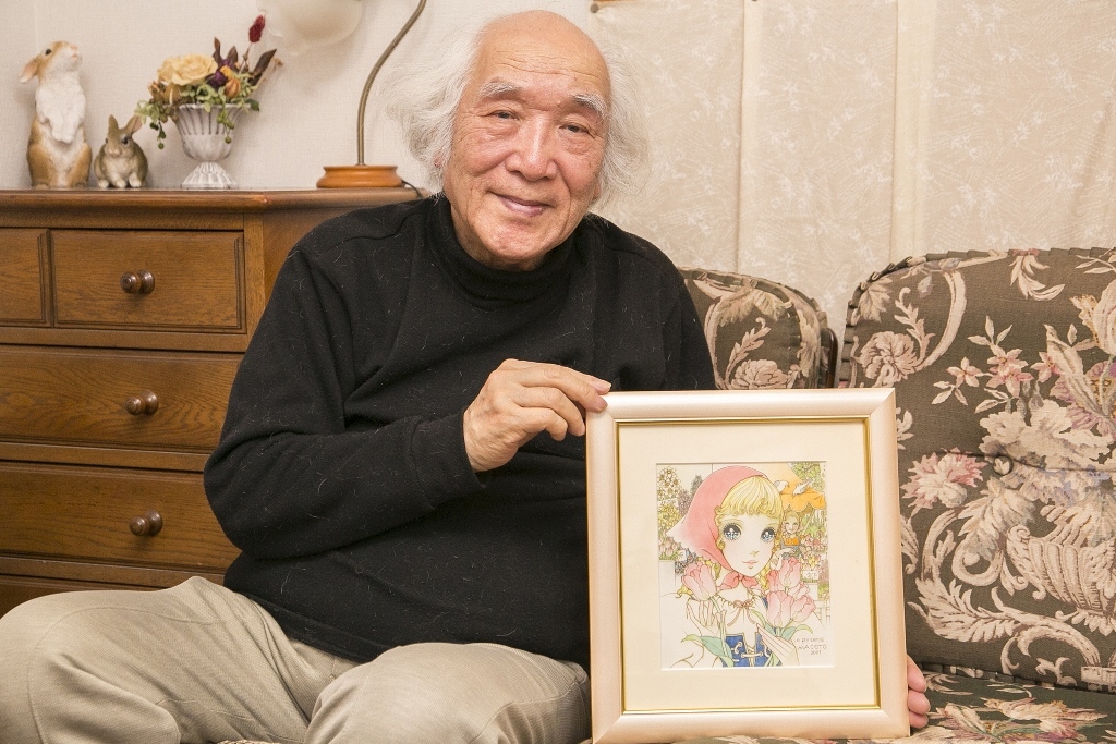 黒いセーターにベージュのズボンを履き、ご自身が書かれた少女漫画の絵を持ってソファー似腰かけている高橋真琴さんの写真