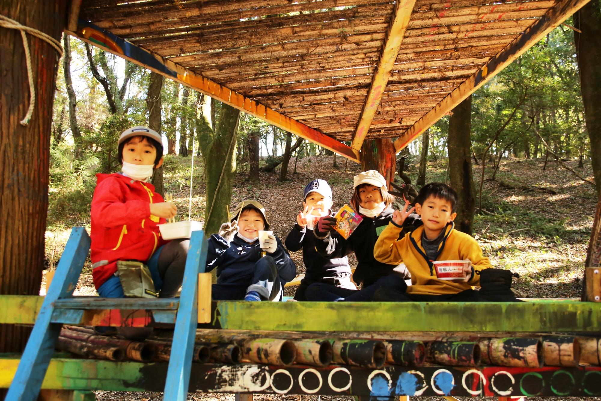 森の中のツリーハウスで子ども達がカップラーメンを食べながらピースをしている写真 魅力発信サイト「サクライク」にリンク
