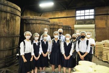大きな樽の置かれた工場内で学生達が工場見学をしている記念写真 魅力発信サイト「サクライク」へリンク