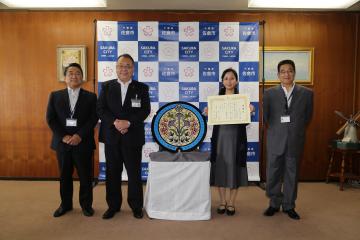 中央に飾られたデザインマンホール蓋、その左側に染井副市長と西田市長、右側に賞状を持った中島さん、関口上下水道事業管理者が並んで立っている写真