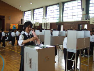 体育館に設置された記載台で生徒達が記載しており、女子生徒が投票用紙を投票箱に入れている写真（中学校生徒会選挙の支援（投票箱の貸出と投票風景））