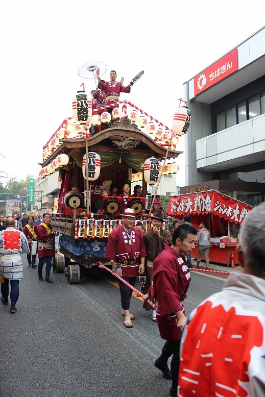 ちばぎんの建物の前の道路を「宮小路町」の神輿がボルドー色の法被を着た人たちにひかれている写真