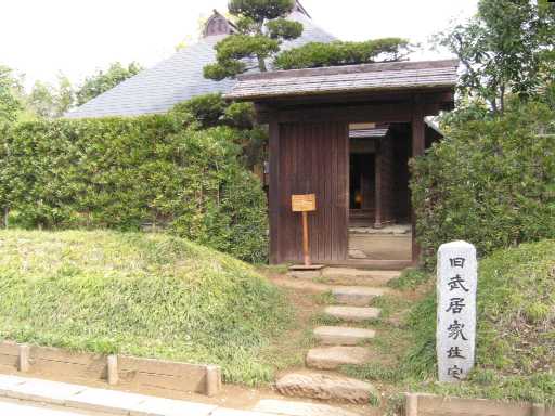 入口右に石碑が建ち、右側の門の奥に旧武居家住宅の母屋が見える写真