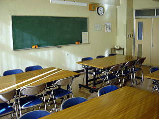 壁に時計と黒板が設置され、パイプ椅子6脚が向い合うように置かれた長机が並んでいる研修室1の写真