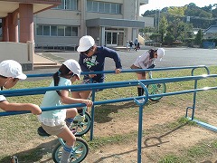 2本並んだ横長の鉄棒で作られた練習用の用具を用いて、両手でその鉄棒を持って身体を支えながら一輪車に乗り練習している数人の生徒たちの様子を横から写した写真