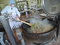 青い帽子の作業員が大きなしゃもじで大きな鍋の蓋を開けてかき回している写真