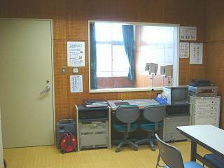 放送機材が置かれている机から窓越しに校内の風景が見える放送室の写真