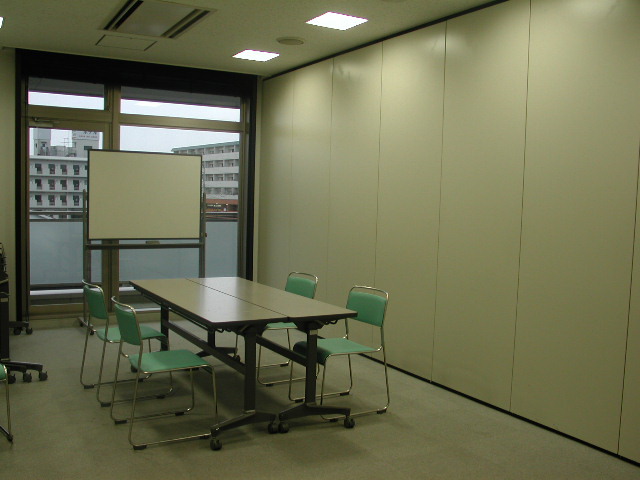 窓側にホワイトボード、中央に2台の長机を組み合わせ、椅子が4脚置かれている第1会議室の写真