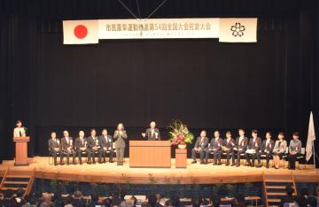 会場の舞台中央の壇上に立ち、話をしている男性、男性の横に立つ手話通訳者、胸に赤い胸章を付けた関係者の方々が並べられたパイプ椅子に座っている佐倉市民音楽ホールでの開会式の写真