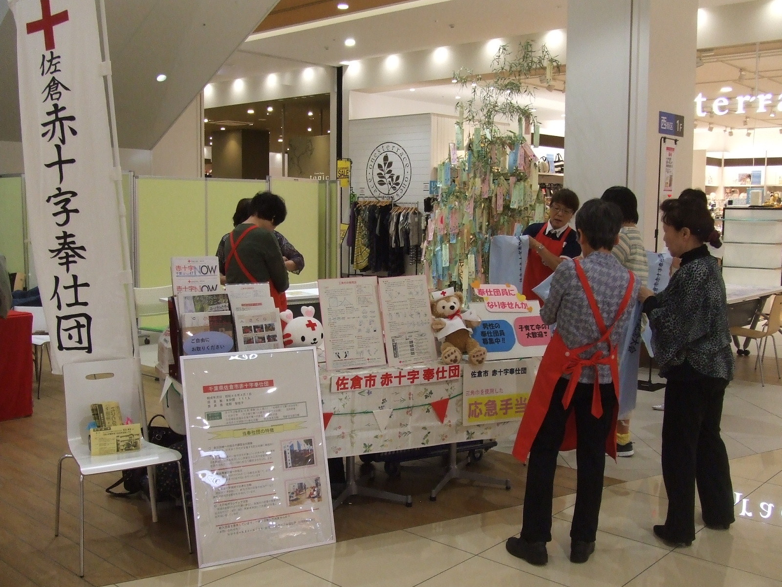 「佐倉赤十字奉仕団」と書かれたのぼりと、展示物などが置かれたブースの周りに人が数人集まっている写真