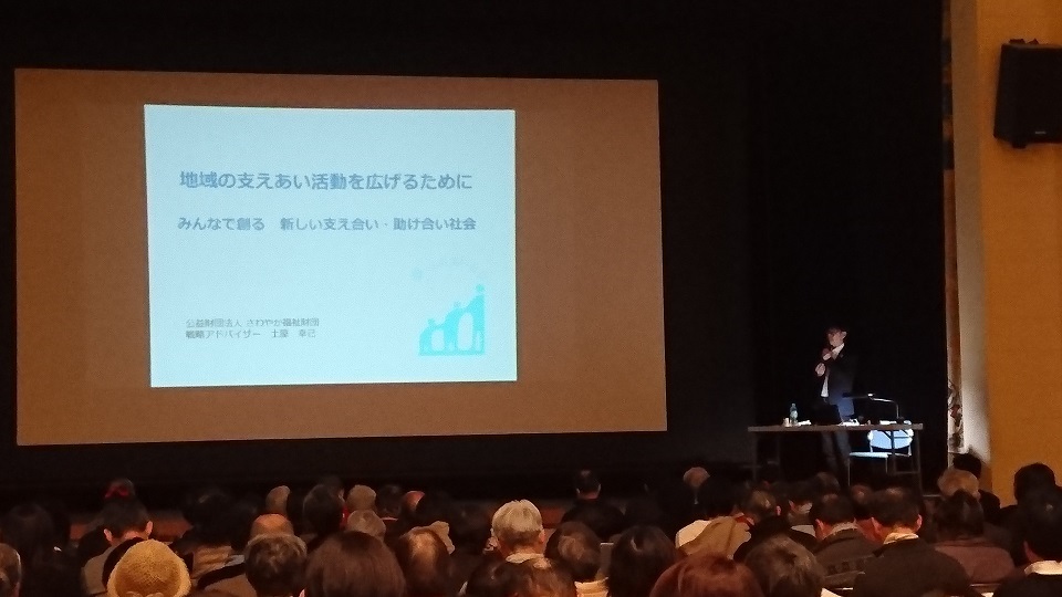 ステージ上でスクリーンに写し出される資料をもとに講演をする土屋 幸己氏と、多くの参加者の方々の写真