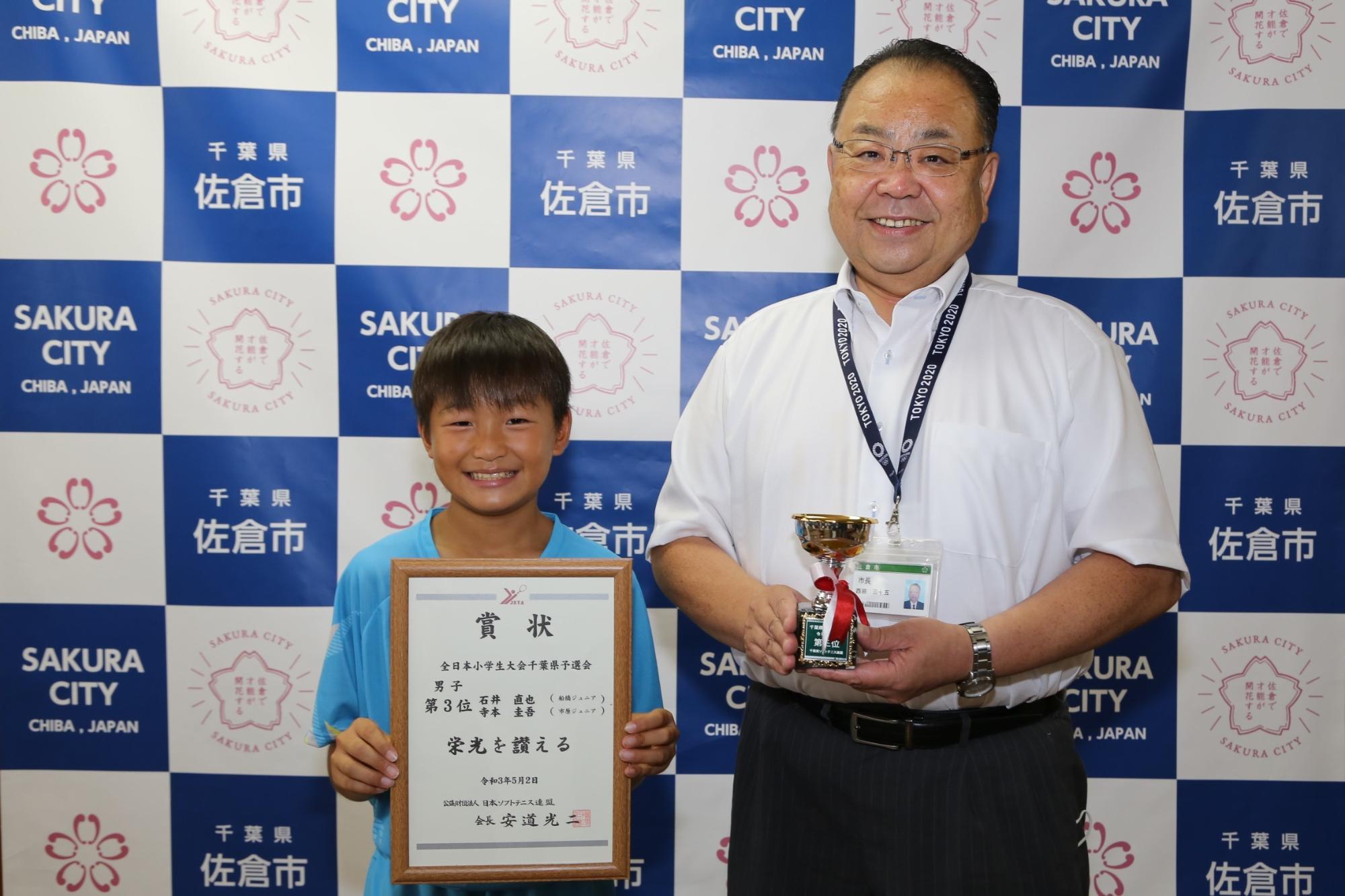 笑顔で額に入った賞状を持つ男の子と、その横でトロフィーを持つ市長の写真
