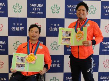 笑顔の男性2名が花の形のメダルを付け、写真が貼られた色紙のようなものを持っている写真