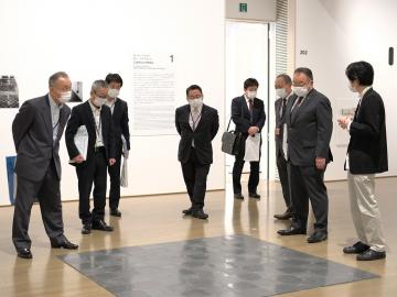 市長や関係者が、床にあるグレーの四角い板のような作品を見ている写真