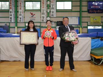 宇山芽紅選手が中央でメダルを持ち、その左右で額入りの賞状を持った女性と、花束を持った市長が並んでいる写真