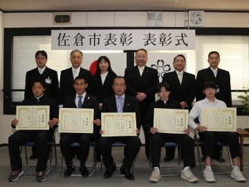 スポーツ功労者表彰式にて、賞状をもって前列に着席している5名の受賞者と、その後ろに並ぶ市長や関係者達が記念撮影をしている写真