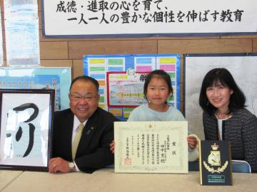 額に入った書道を持つ市長、賞状を持った女子生徒、表彰楯の後ろの女性が並んで記念撮影をしている写真