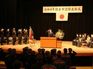 舞台上の左側に並ぶ正装した消防団員と、演台に立つ男性が敬礼をし、右側に関係者達が着席している写真