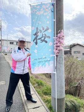 桜の漢字が書かれ、桜の花びらが描かれた旗の端を持って立っている市長の写真