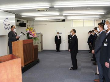 左側の演台で話をしている市長と、右側にスーツを着た職員が横一列に並び、その前に男性1人が立っている写真