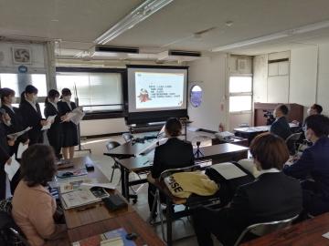 プロジェクタースクリーンに映る資料を注目する学生や関係者と、左側に立っている男女学生の写真