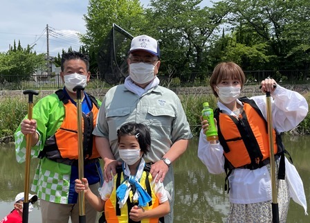 緑の法被を着た男性、子供、岡田ロビン翔子さんがオールのようなものを持って、市長を中心に集まっている写真