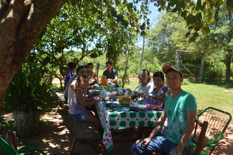晴れた日の木の陰でテーブルに座り現地の方たちと食事会をしている稲葉健一さんの写真