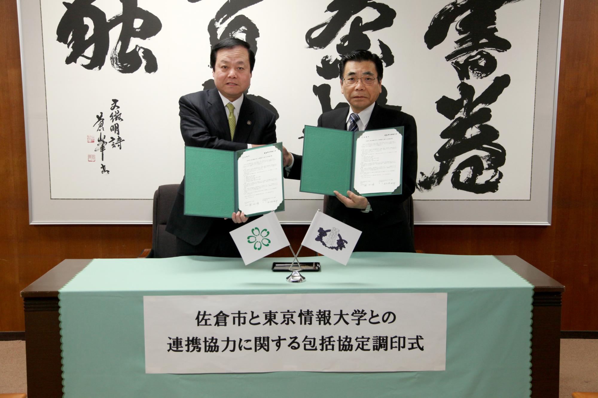 緑色のホルダーに協定書があり、そのホルダーを蕨市長と東京情報大学 牛久保学長がお互いに持ち握手を交わしている写真