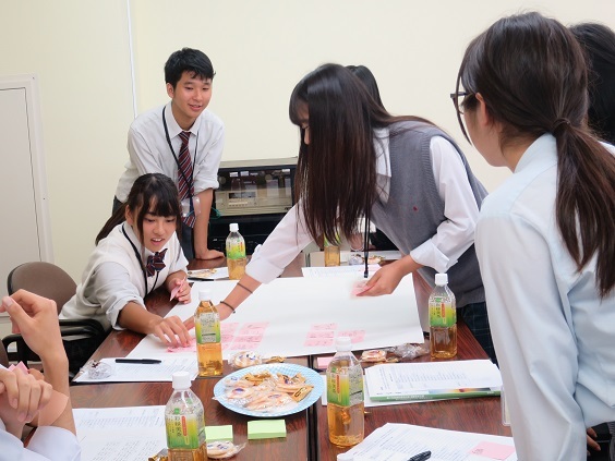 模造紙に書き込みがされた付箋を貼り付けている女子学生と、周りでその様子を見ている男女学生の写真