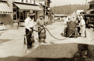 2名の男性がホースの先を持ち、奥の機械を数名の人達が操作している道路舗装工事の白黒写真