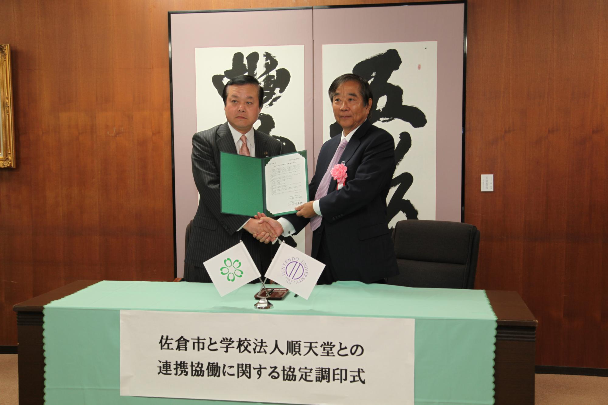 書物が記載された屏風の前で協定書を手に握手を交わしている蕨市長と順天堂理事長 小川さんの写真