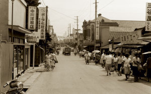 道路の両端に昭和初期の看板の店が並ぶ新町商店街で買い物をしている人達を写した白黒写真