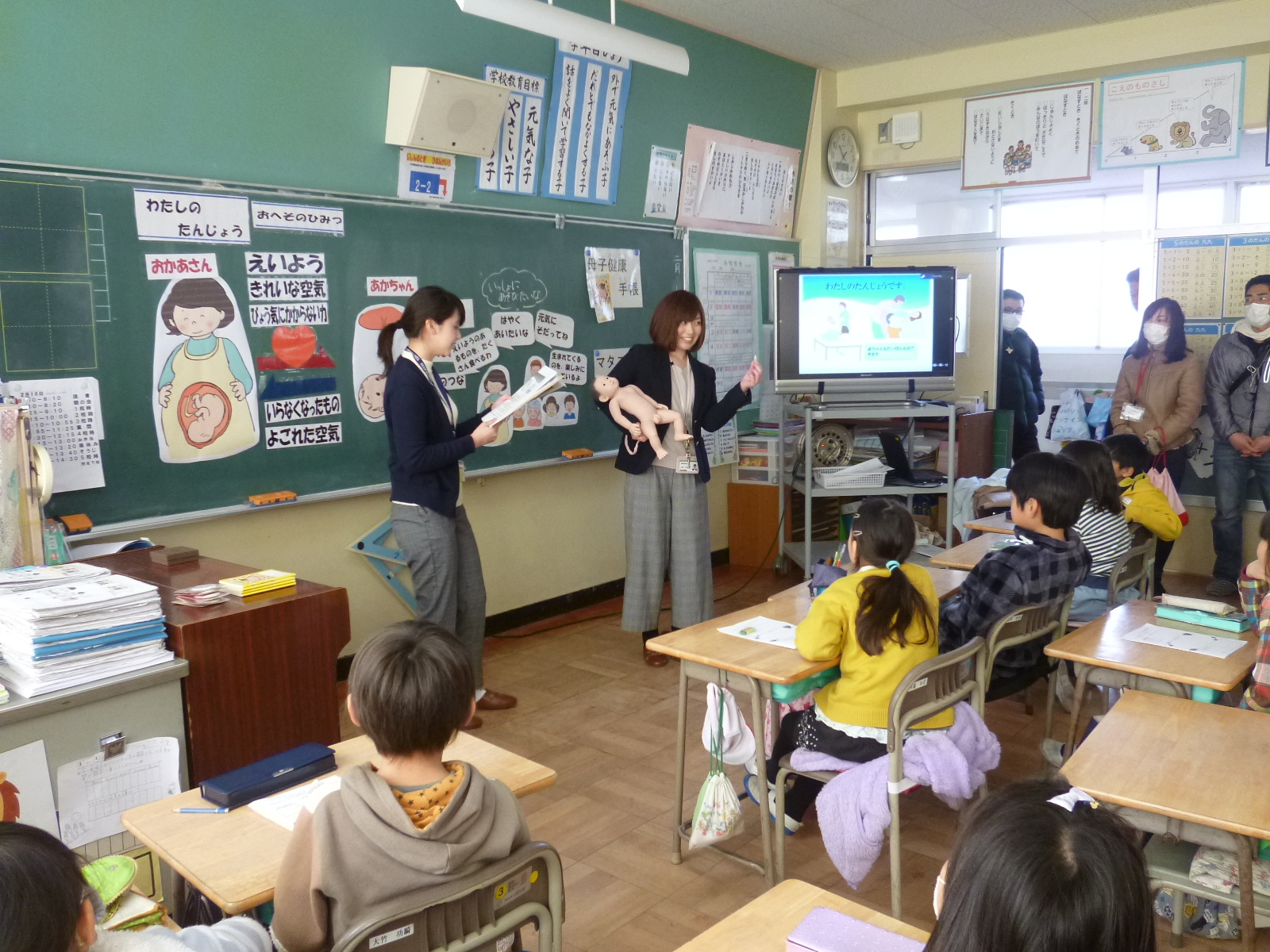 黒板の前に立って説明している先生と赤ちゃんの人形を抱いている地区担当保健師、授業を受けている生徒たちの写真