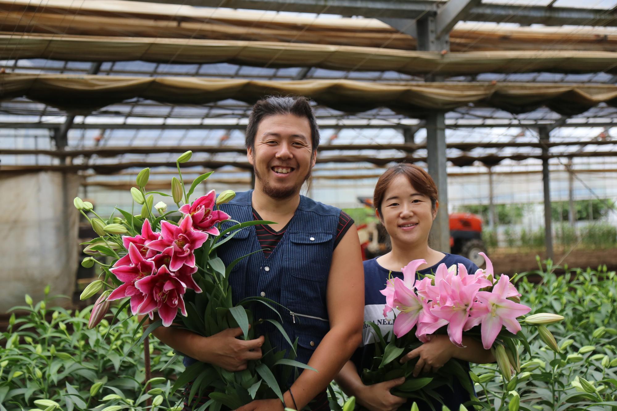 ユリを育てているハウスの中でピンクのユリの花を持っている笑顔の山崎さんご夫妻の写真