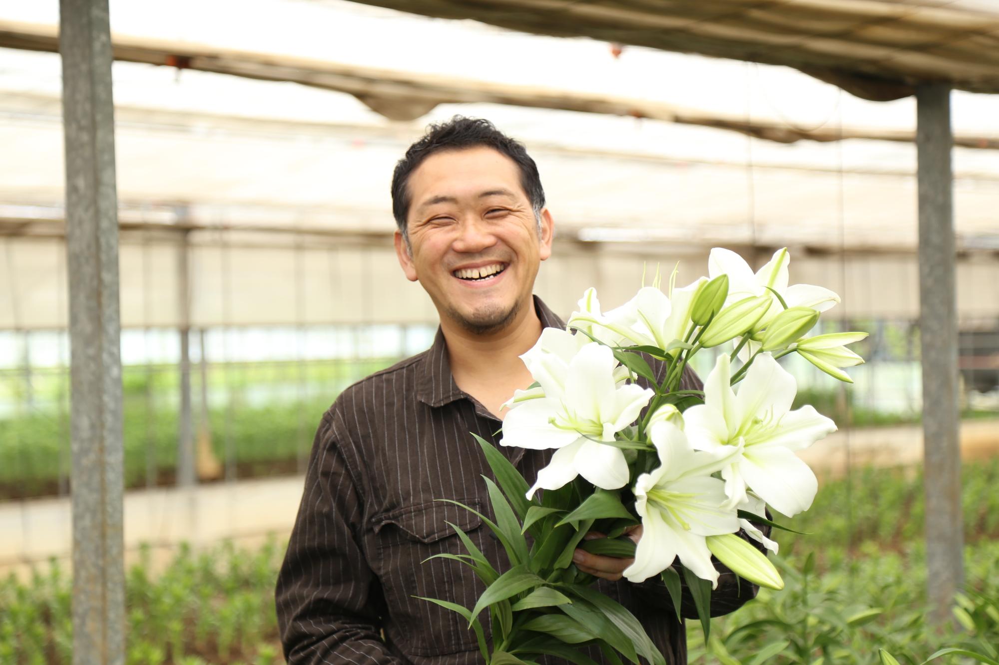 百合を育てているビニールハウスの中で白いユリの花を持っている笑顔の男性の写真