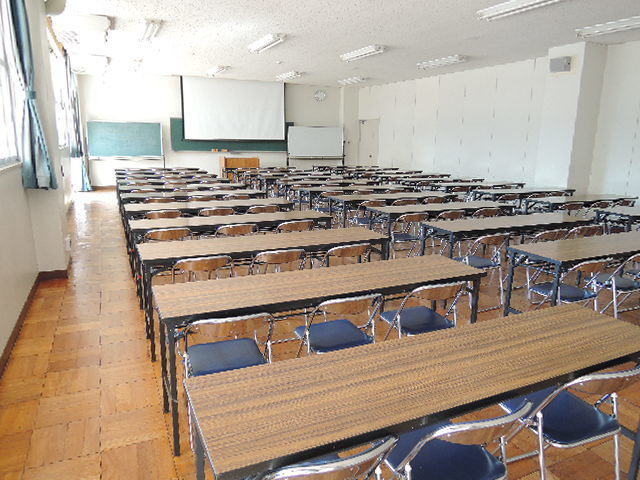 奥に黒板とスクリーンがあり、パイプ椅子が3脚ずつセットされている長机が2列ずつ並んでいる研修室の写真