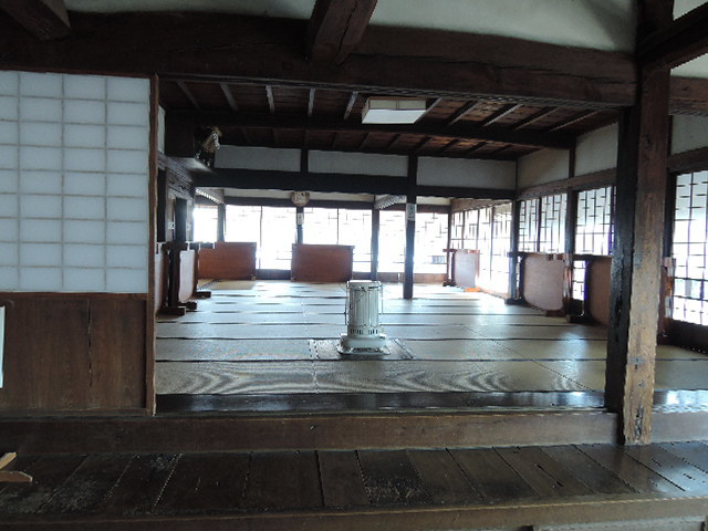 6つのテーブルが脇に立てかけられ、中央にストーブが設置されている和室の室内の写真