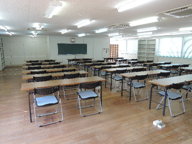 奥に黒板があり、2脚ずつパイプ椅子がセットされている長机が設置されている実習室の写真