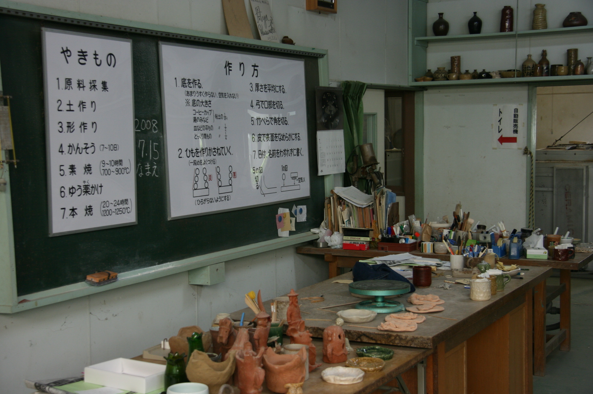 黒板にやきものと作り方が大きな模造紙で貼られて、手前の作業台の上に道具などが置いてある陶芸教室の写真