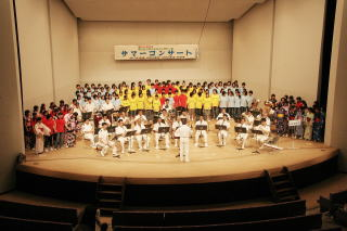 舞台の上に白や赤・黄色の衣装を纏ったコーラス隊の前で打楽器を演奏しているサマーコンサートの写真