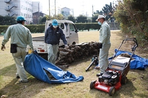 軽トラックの横に積まれた木材、芝刈り機と清掃作業をしている3人の職員の写真