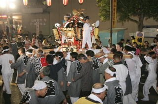 大きな神輿にこどもが団扇を手に持って立っており、その神輿をたくさんの法被を着た大人が担いでいる写真
