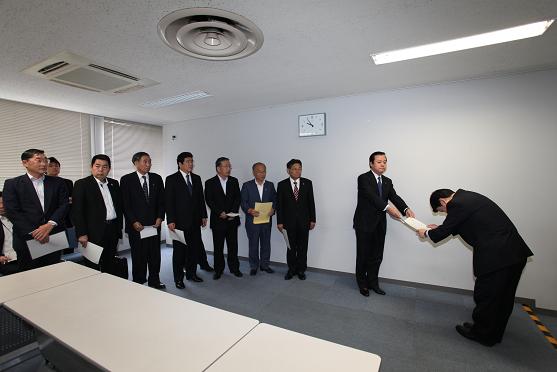 8名のスーツを着た人たちが見守る中、男性がお辞儀をしながら書類を関係者の男性に渡している写真