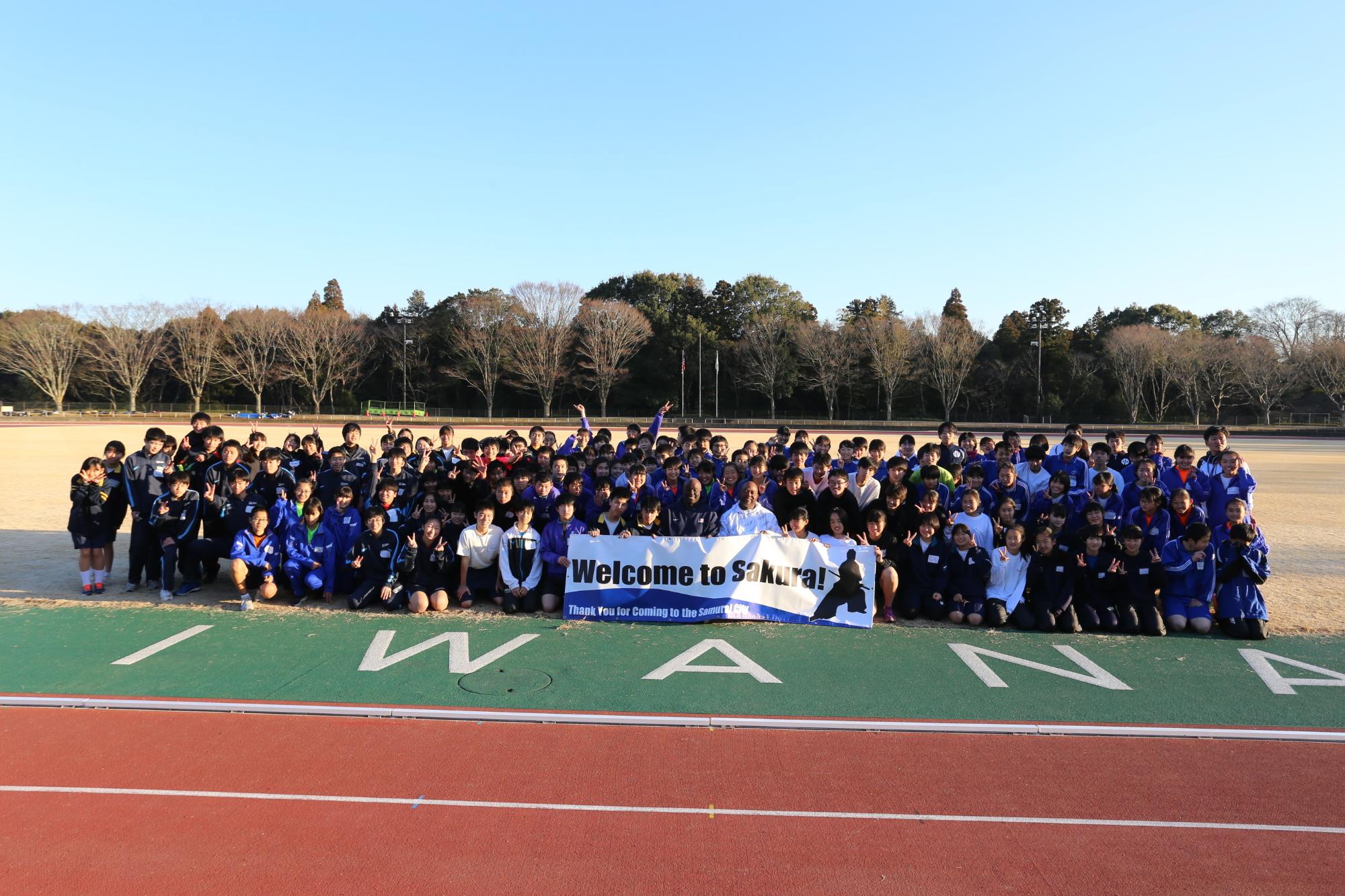 「Welcome to Sakura！」と書かれた横断幕を前に、陸上クリニックの参加者皆さんとアメリカ陸上競技連盟所属コーチの方々が集まって写っている集合写真