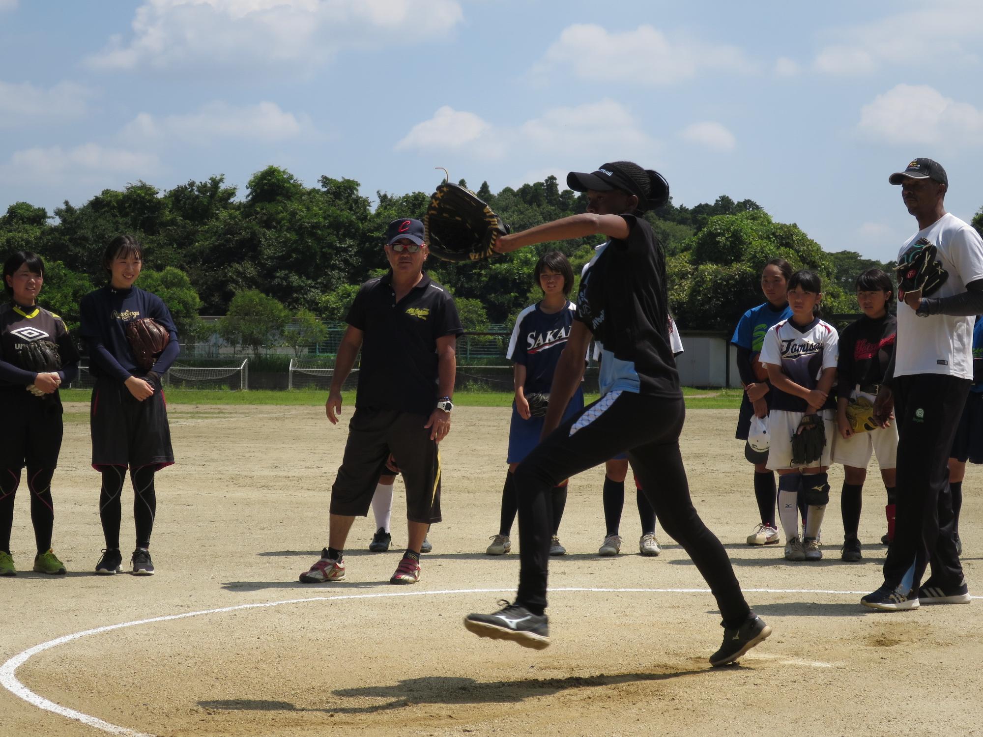 左手にグローブを付けたボツワナの選手がボールを投げようとしており、生徒達が近くで選手が投げる様子を見ている写真